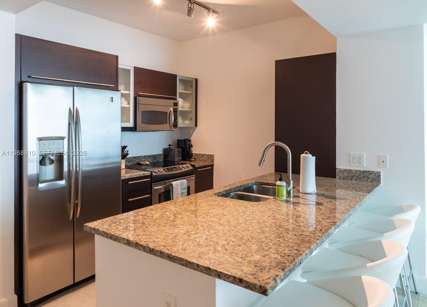 A kitchen features granite countertops, stainless steel appliances, dark wood cabinets, and white bar stools — 950 Brickell Bay Dr # 1504, Miami FL 33131, MIAMI, FL