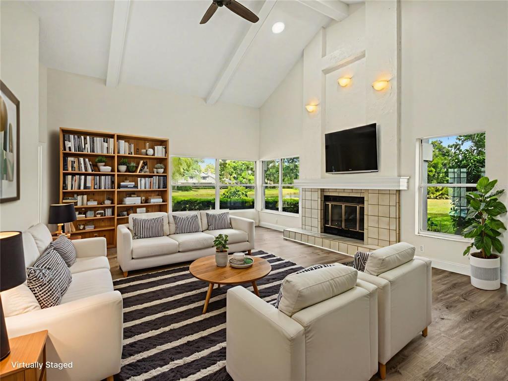 A living room with a vaulted ceiling, fireplace, bookshelves, and large windows overlooking greenery — 7505 WEEPING WILLOW BOULEVARD, SARASOTA, FL
