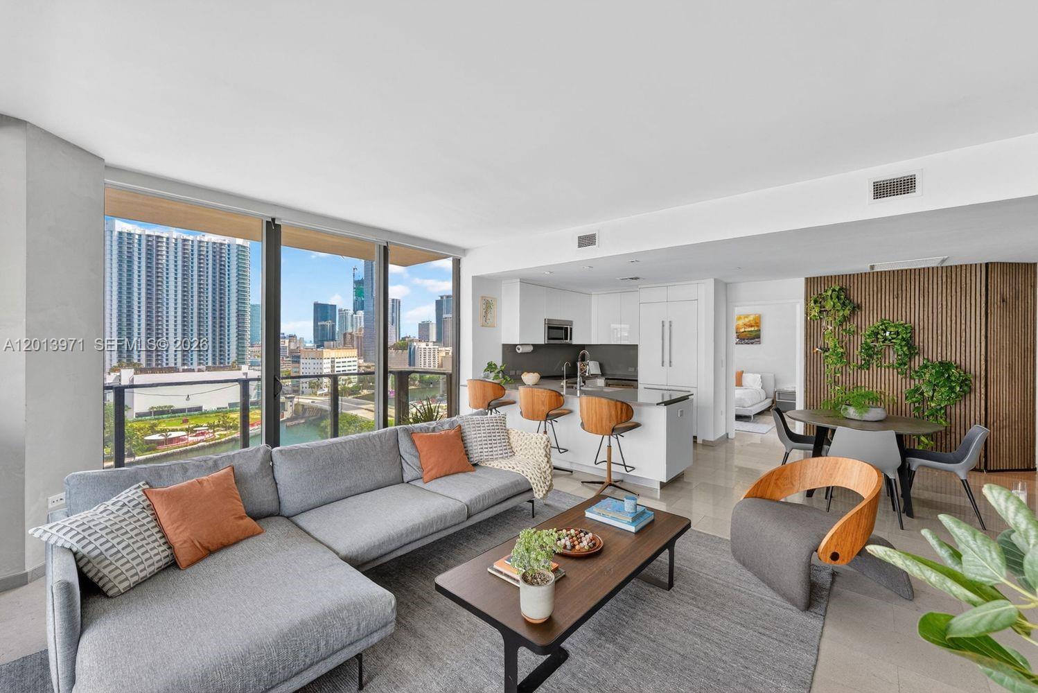 A modern living room with a sectional sofa, kitchen island, and city views through floor-to-ceiling windows — 88 SW 7th St # 1012, Miami FL 33130, MIAMI, FL