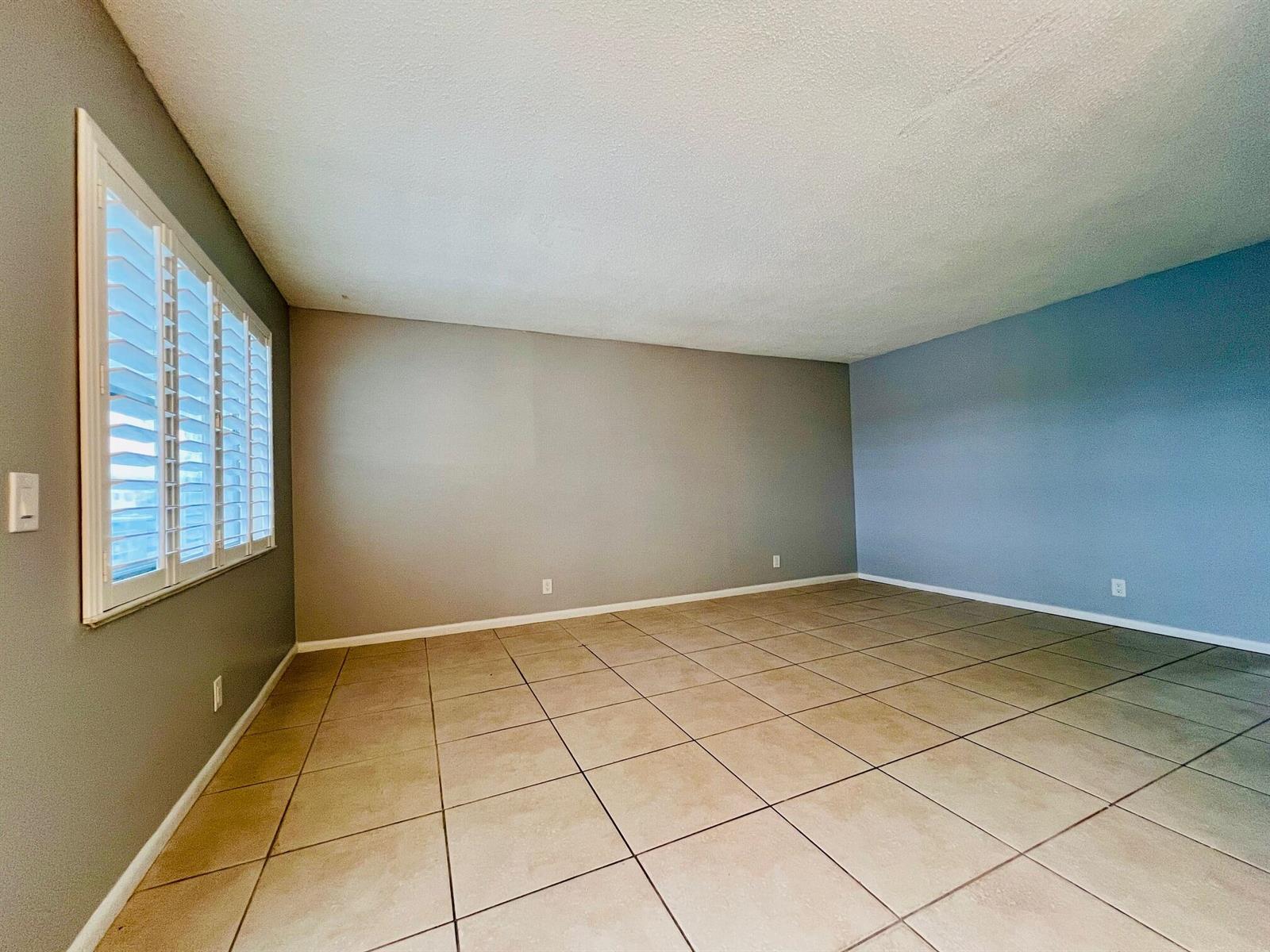 An empty room with beige tile flooring, gray and blue walls, and white plantation shutters — 74 Berkshire C, West Palm Beach FL 33417, WEST PALM BEACH, FL