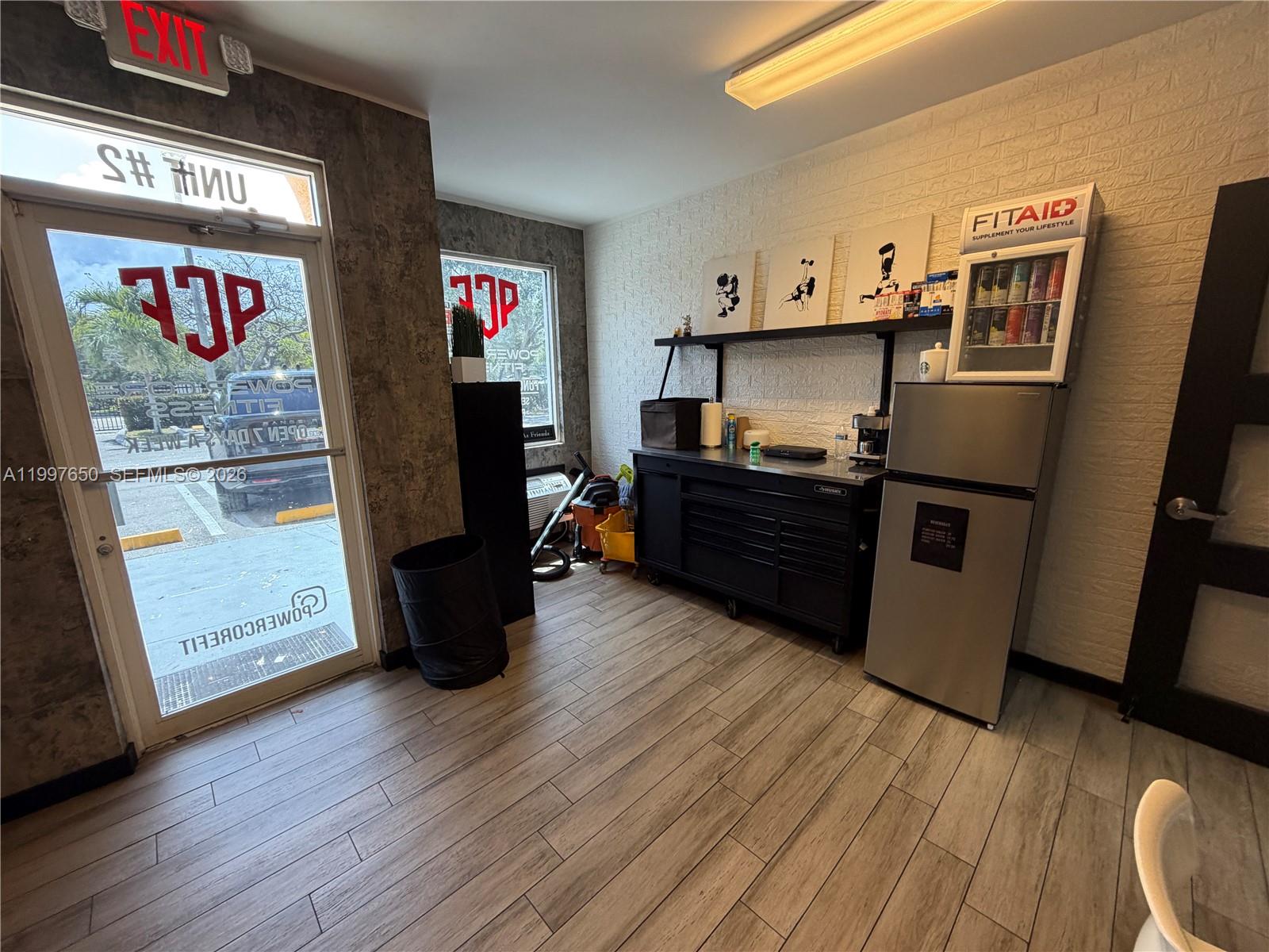 A small gym reception area features wood-look tile flooring, a black workbench, and a refrigerator — 14911 SW 137th St # 14911, FL 33196, FL
