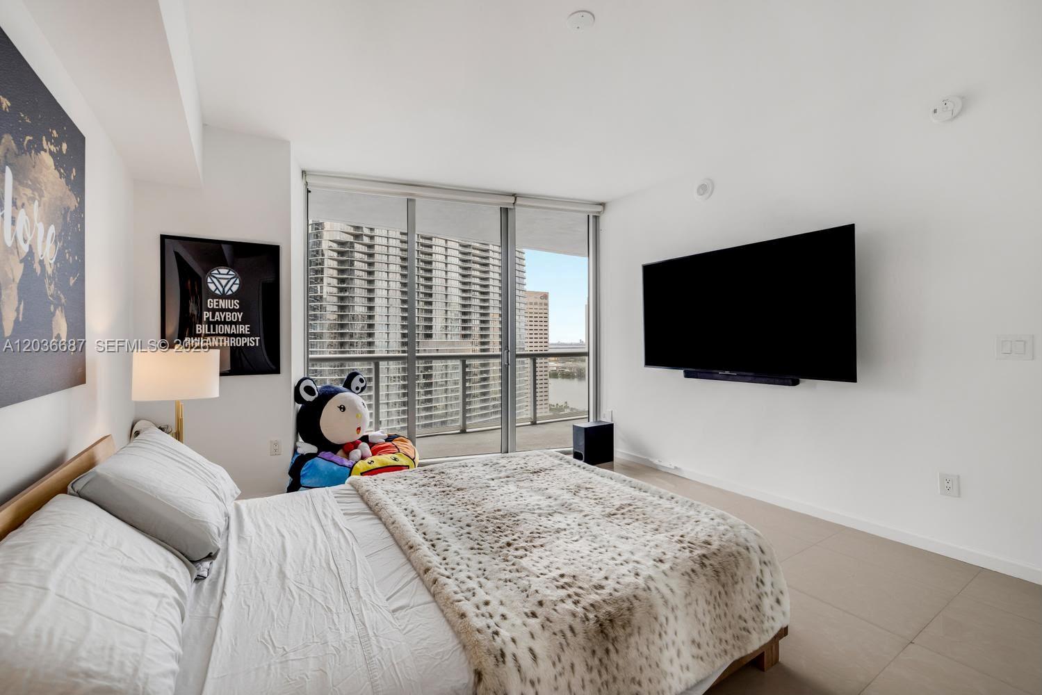 A bedroom features a bed with a patterned throw, a wall-mounted television, and floor-to-ceiling windows — 475 Brickell Ave # 3214, Miami FL 33131, MIAMI, FL