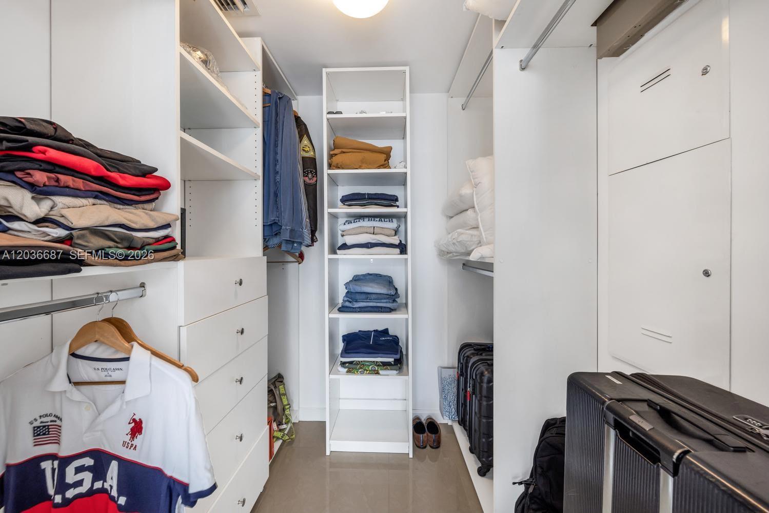A walk-in closet features white shelving, hanging clothes, folded garments, and luggage on the floor — 475 Brickell Ave # 3214, Miami FL 33131, MIAMI, FL