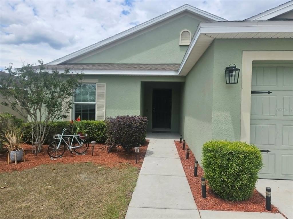 A light green house exterior features a concrete walkway leading to a dark front door — 30939 WATER LILY DRIVE, BROOKSVILLE, FL