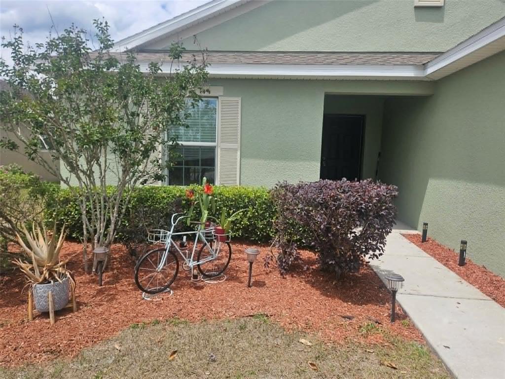 A light green house exterior features a front walkway, landscaping, and a decorative bicycle — 30939 WATER LILY DRIVE, BROOKSVILLE, FL