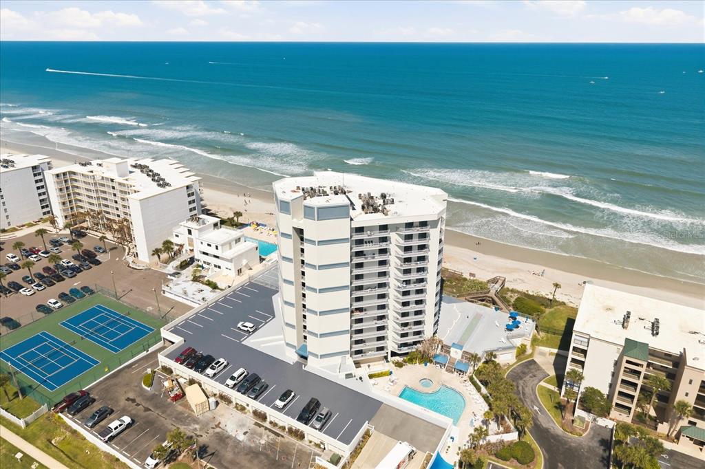 An aerial view shows a high-rise beachfront condominium with a pool, tennis courts, and parking — 5207 S ATLANTIC AVENUE UNIT 1025, NEW SMYRNA BEACH, FL