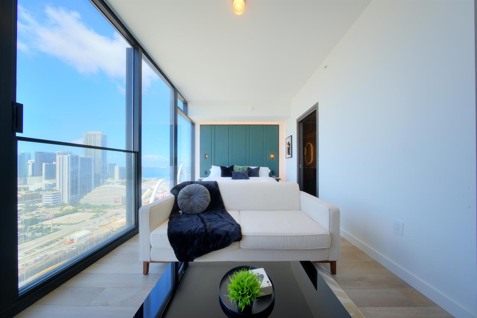 A modern bedroom features a white sofa, a bed with a green headboard, and floor-to-ceiling windows — 20 NE 11th Street # 3205, Miami FL 33132, MIAMI, FL