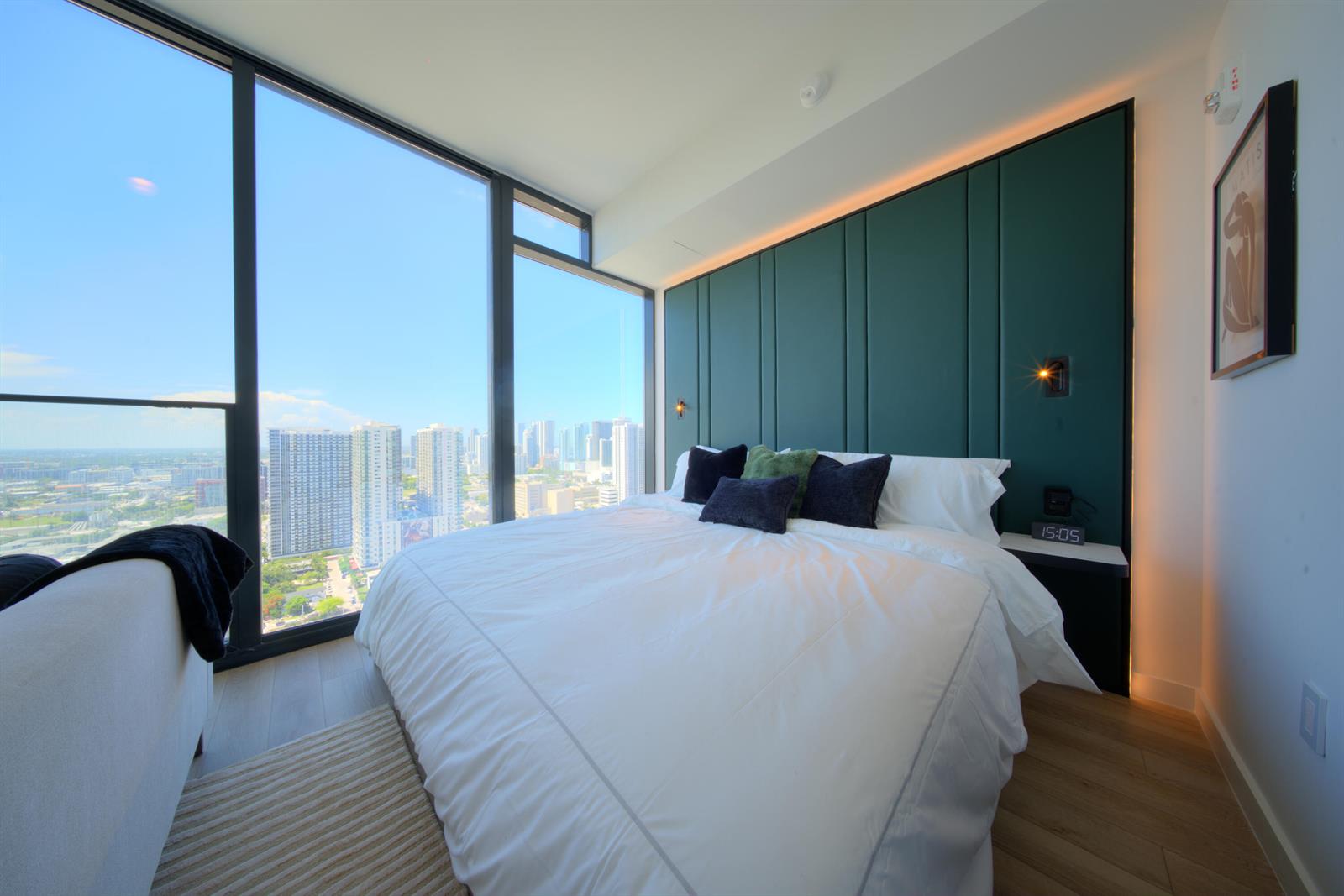 A bedroom with a green paneled headboard, white bedding, and floor-to-ceiling windows overlooking a city — 20 NE 11th Street # 3205, Miami FL 33132, MIAMI, FL