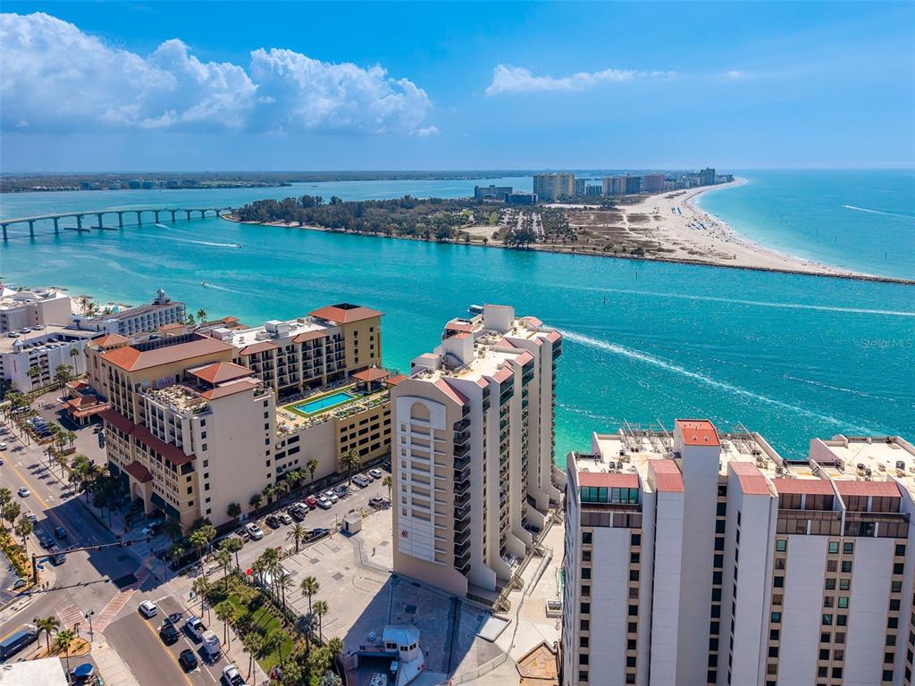 An aerial view shows high-rise buildings near a bridge, a waterway, and a sandy beach — 450 S GULFVIEW BOULEVARD UNIT 208, CLEARWATER BEACH, FL