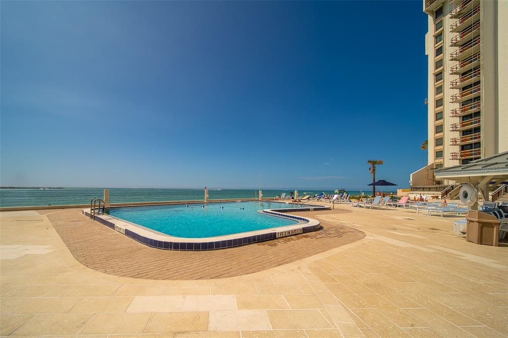 An outdoor swimming pool overlooks the ocean next to a tall multi-story building — 450 S GULFVIEW BOULEVARD UNIT 208, CLEARWATER BEACH, FL