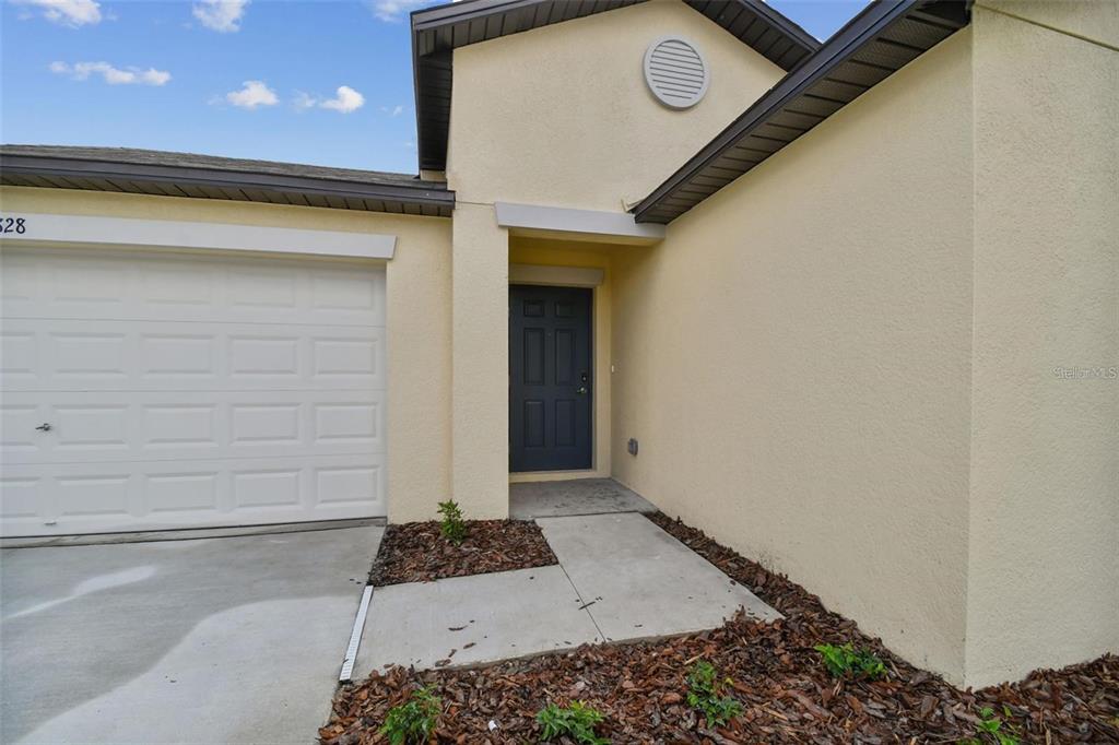 A beige house exterior features a white garage door, a dark front door, and landscaping — 9828 BRANCHING SHIP TRACE, WESLEY CHAPEL, FL