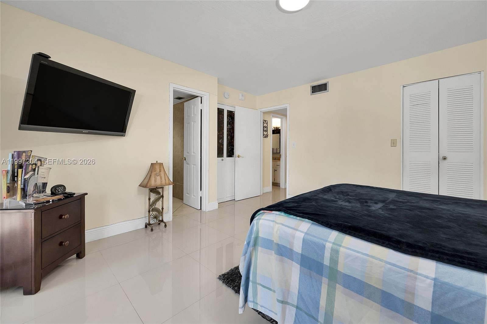 A bedroom features light-colored walls, tiled flooring, a mounted television, and multiple closet doors — 7620 Carlyle Ave # 204, Miami Beach FL 33141, MIAMI BEACH, FL