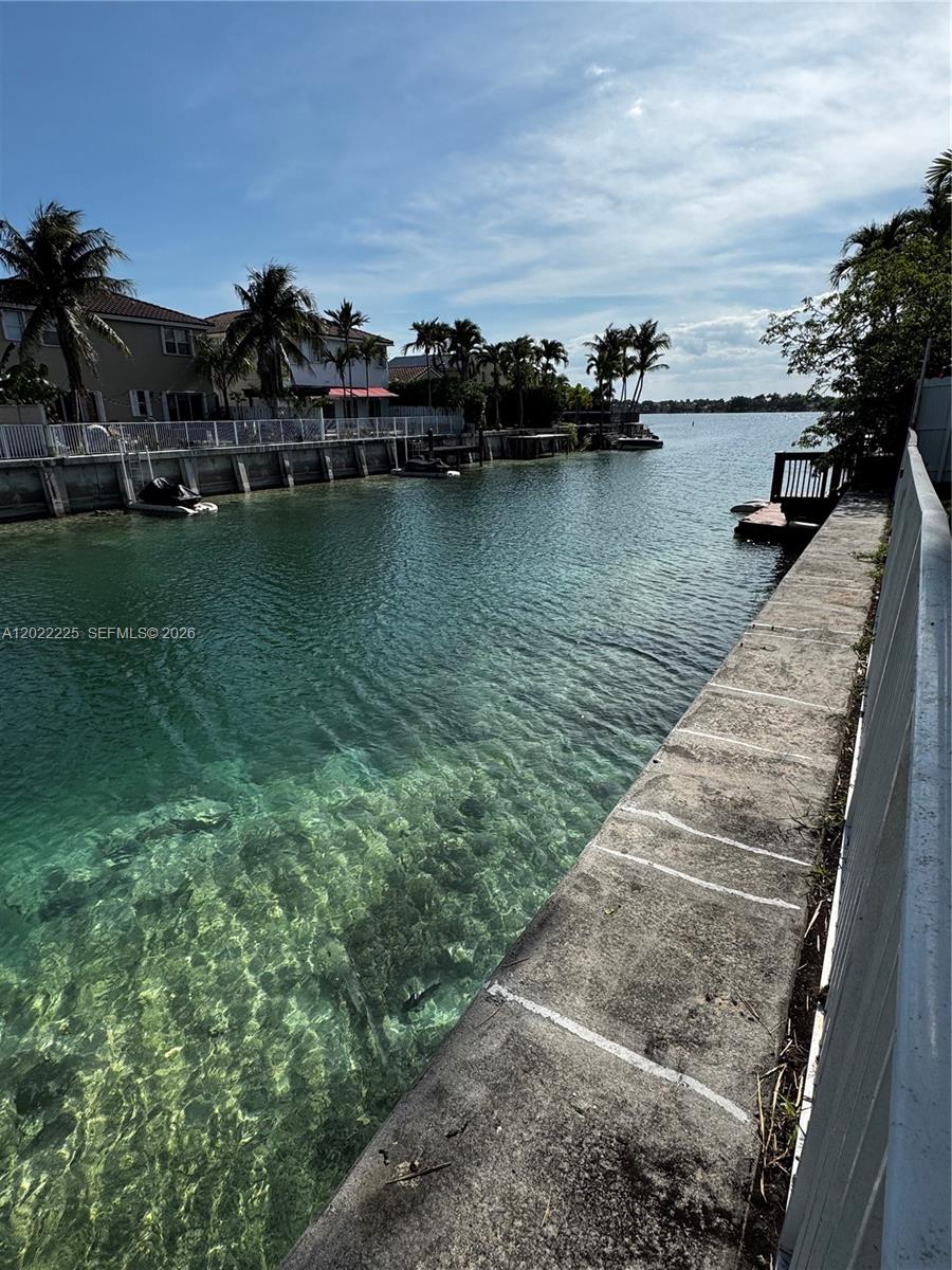 A concrete seawall borders a clear canal with residential homes and palm trees in the background — 13312 SW 144th Ter, Miami FL 33186, MIAMI, FL