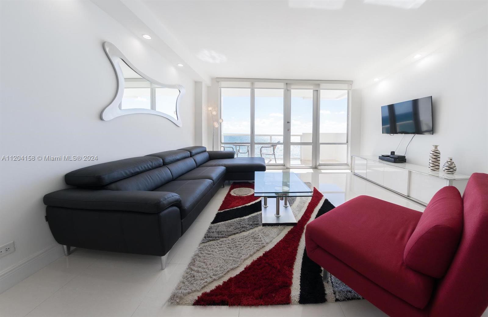 A modern living room features a black sectional, red chair, glass coffee table, and ocean views — 5601 Collins Ave # 1603, Miami Beach FL 33140, MIAMI BEACH, FL
