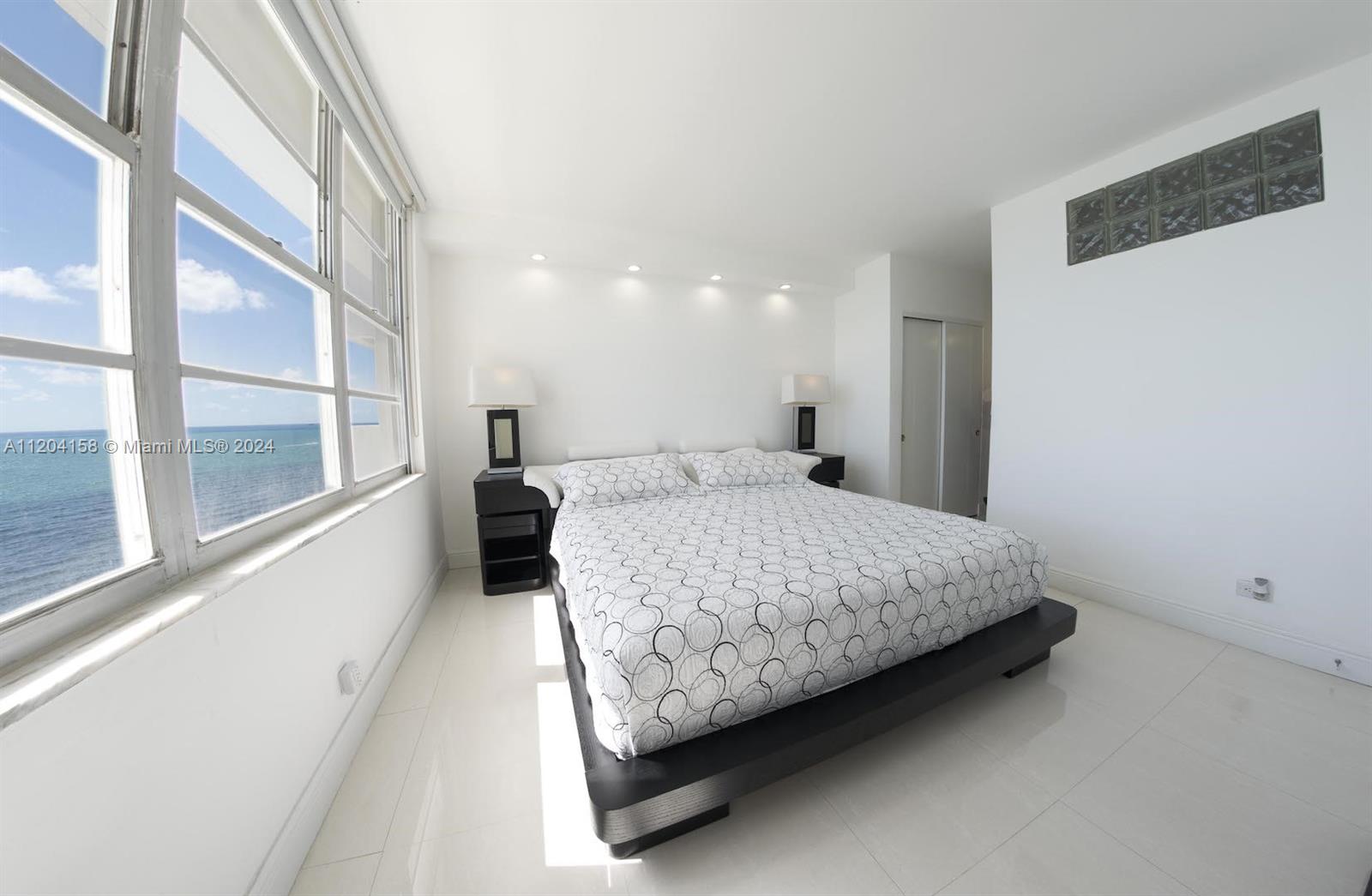A bedroom features a platform bed, white tile floors, and a large window overlooking the ocean — 5601 Collins Ave # 1603, Miami Beach FL 33140, MIAMI BEACH, FL