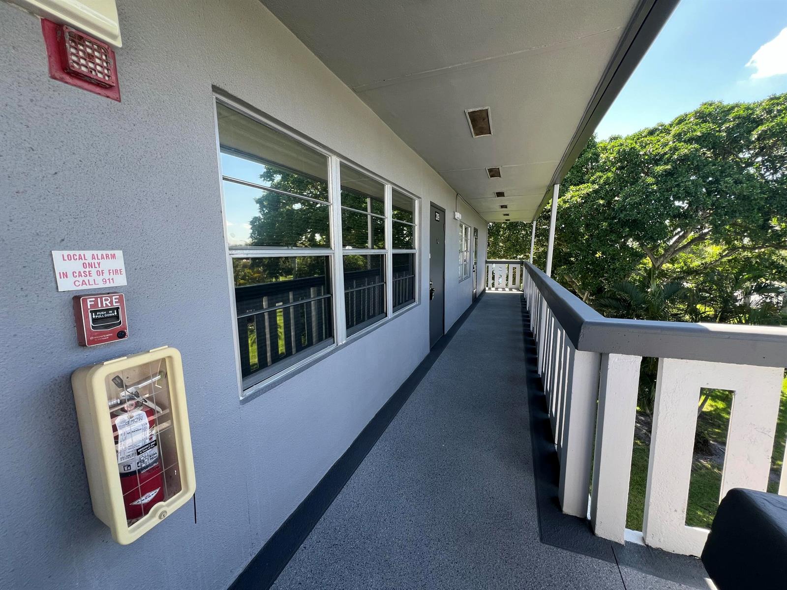 An outdoor apartment walkway features a fire alarm, extinguisher, and a railing overlooking trees — 209 Suffolk E, Boca Raton FL 33434, BOCA RATON, FL