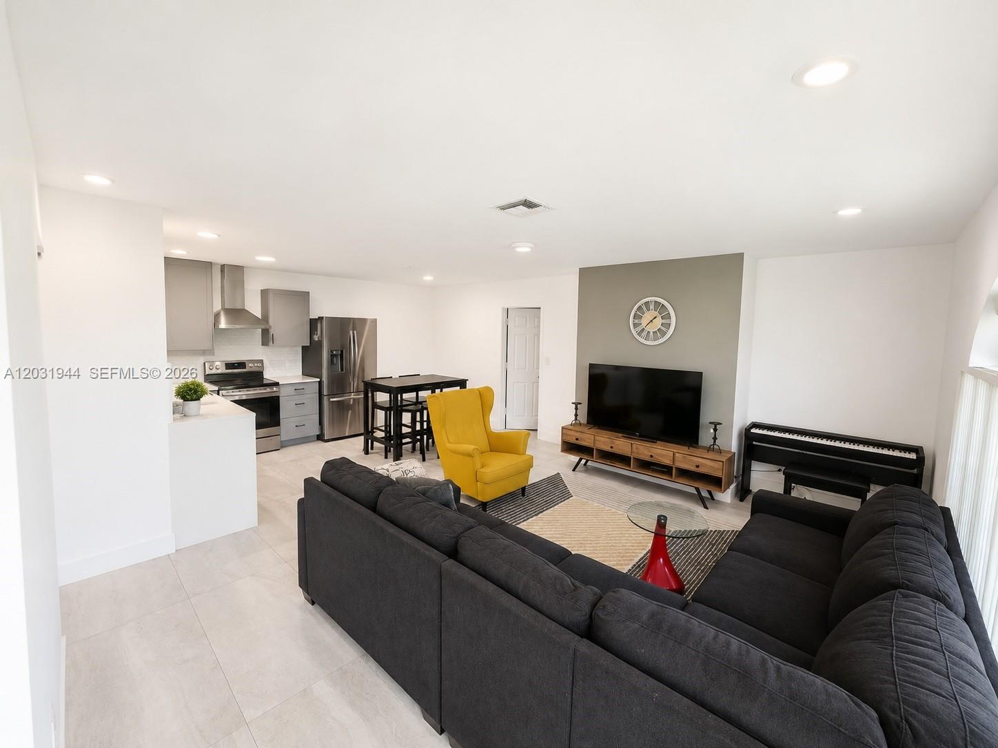 An open-concept living room and kitchen featuring a grey sectional, yellow armchair, and stainless appliances — 800 NE 2nd Ct, Hallandale Beach FL 33009, HALLANDALE BEACH, FL