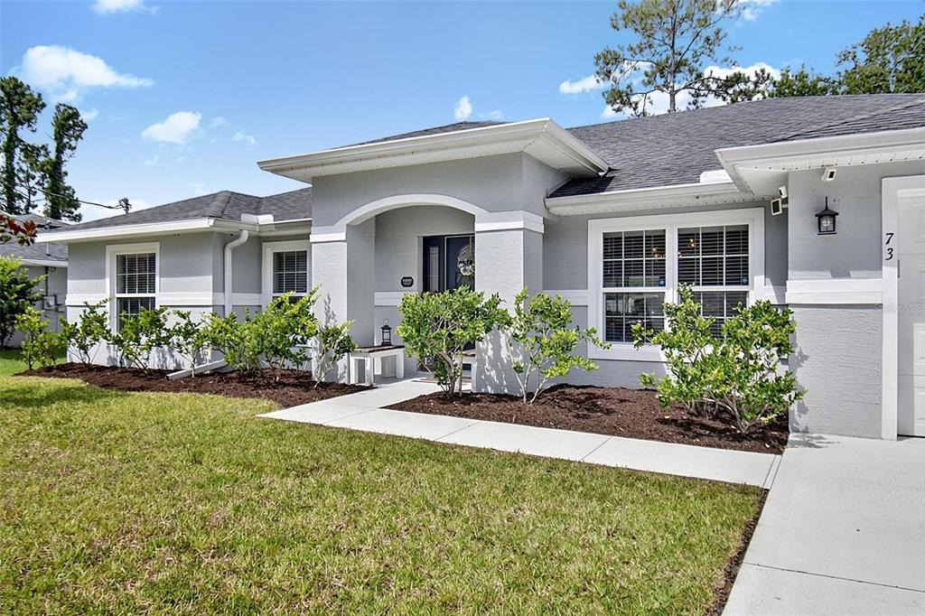 A gray single-story house features a covered entryway, white trim, and landscaped front garden beds — 73 LINDSAY DRIVE, PALM COAST, FL