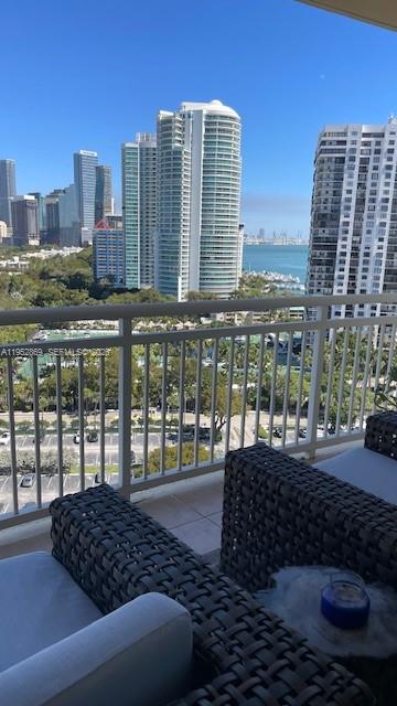A balcony with wicker furniture overlooks a city skyline, a park, and the ocean — 2475 Brickell Ave # 1704, Miami FL 33129, MIAMI, FL