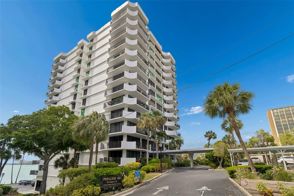 A multi-story white residential building with curved balconies stands next to a paved driveway — 80 ROGERS STREET UNIT 6C, CLEARWATER, FL