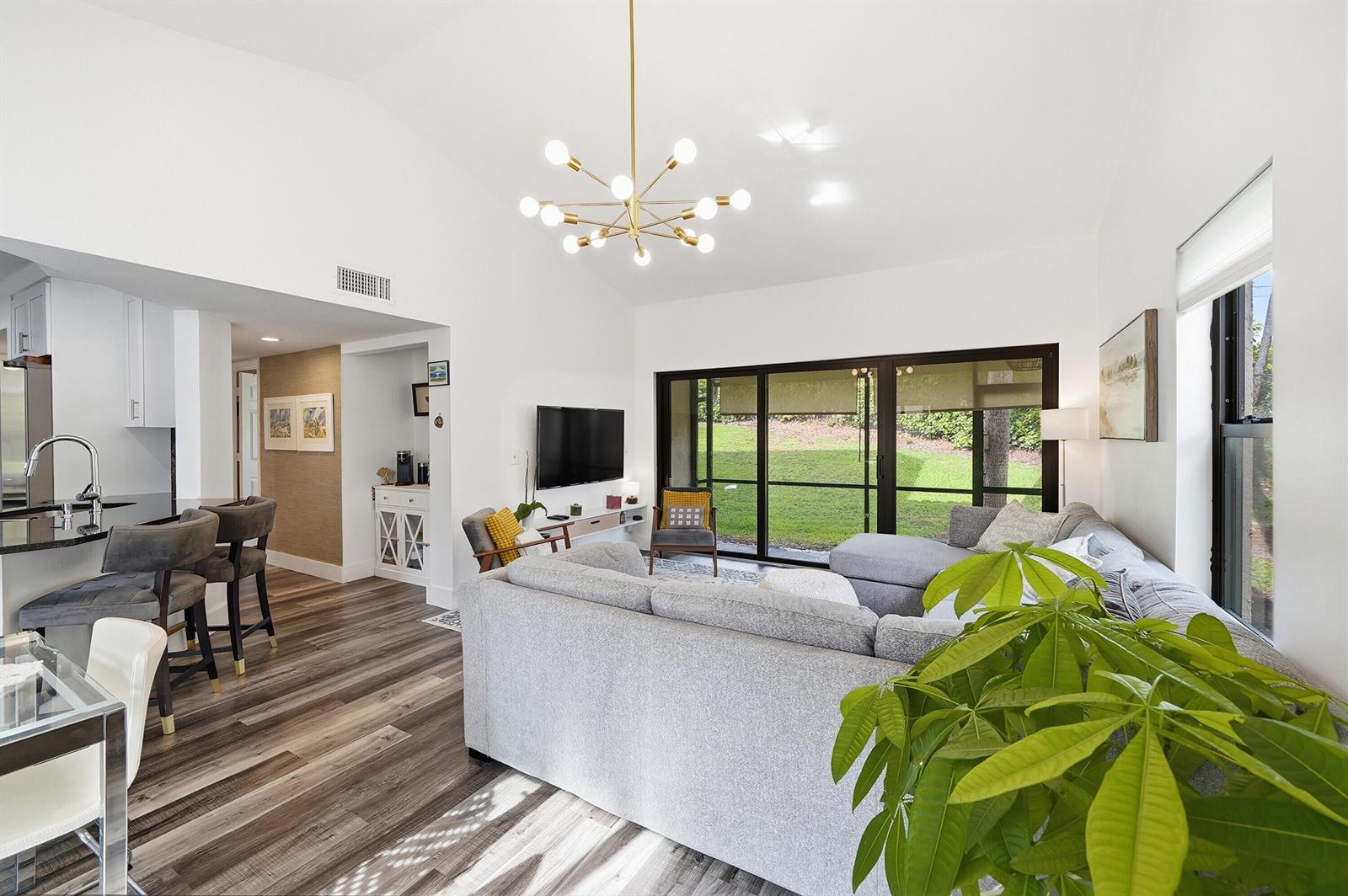 A living room with a grey sectional, modern chandelier, and sliding glass doors to a yard — 5 Via De Casas Sur # 105, Boynton Beach FL 33426, BOYNTON BEACH, FL