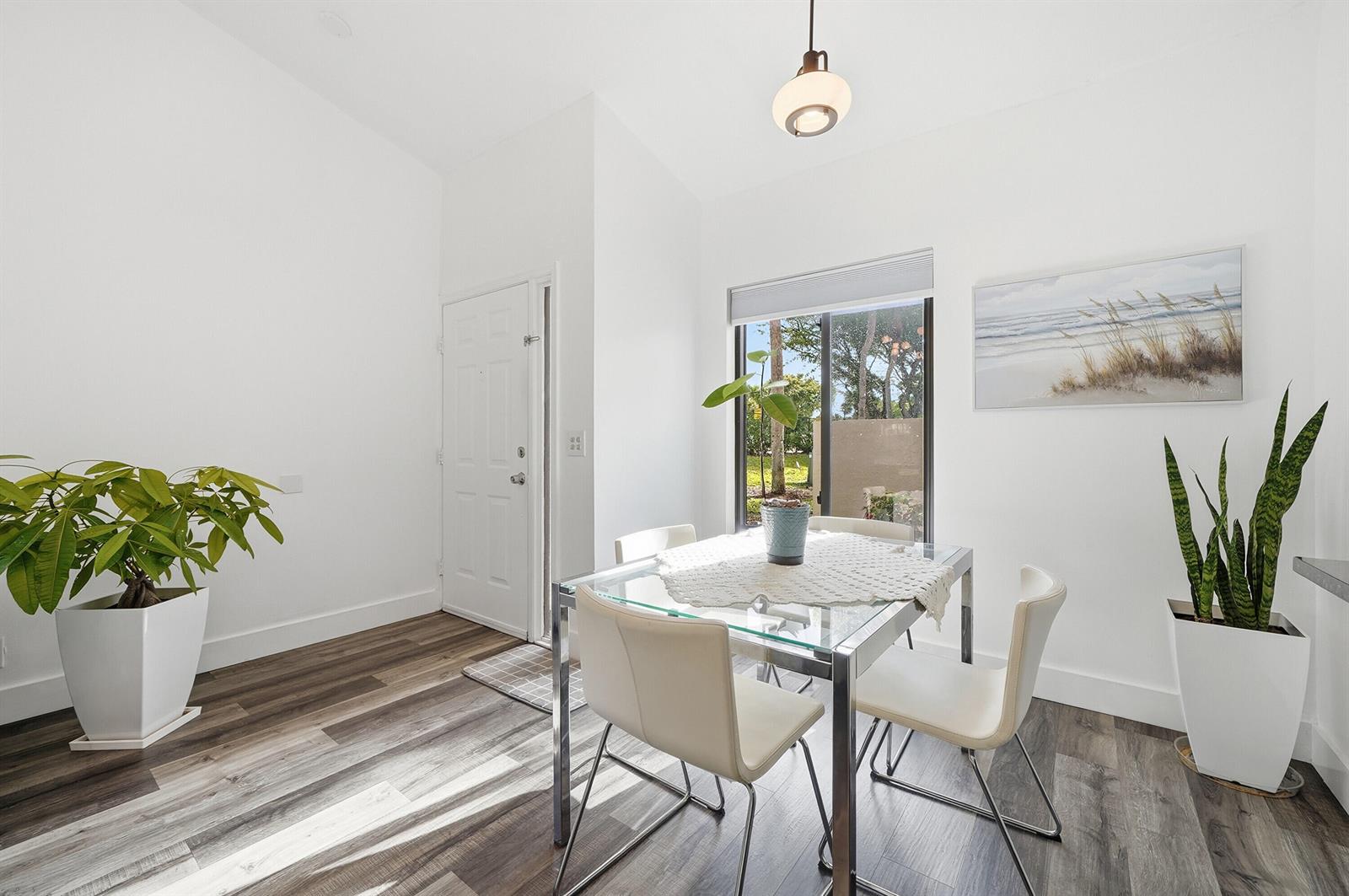 A dining area features a glass table, four white chairs, two potted plants, and wood-look flooring — 5 Via De Casas Sur # 105, Boynton Beach FL 33426, BOYNTON BEACH, FL