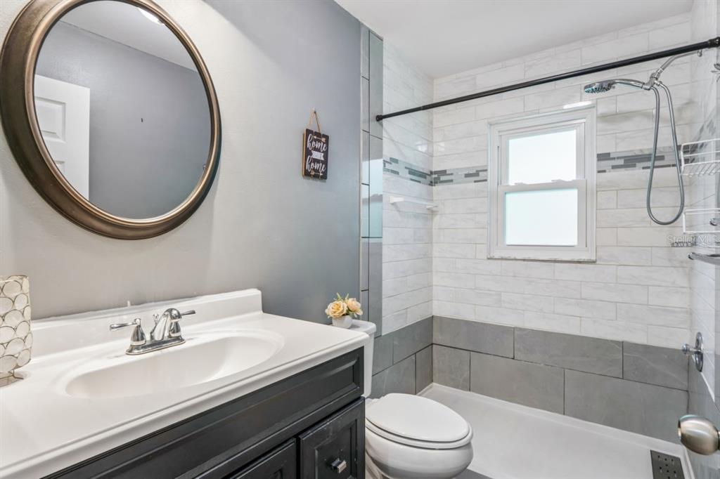A bathroom features a dark vanity, a round mirror, and a tiled shower-tub combination — 6016 WILDS DR, NEW PORT RICHEY, FL