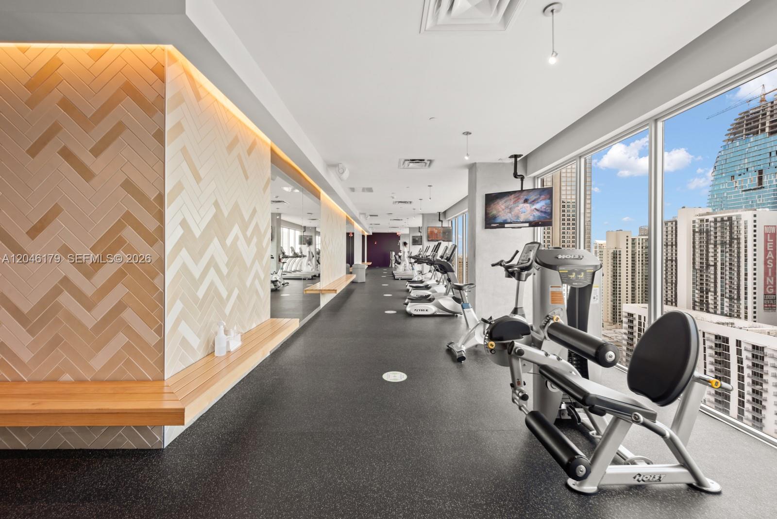 A modern gym features exercise equipment, a herringbone-tiled wall, and floor-to-ceiling windows overlooking city buildings — 151 SE 1st St, Miami FL 33131, MIAMI, FL