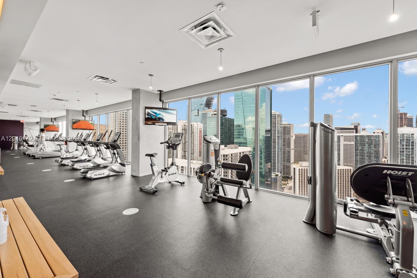A modern gym features exercise equipment, black rubber flooring, and floor-to-ceiling windows overlooking a city — 151 SE 1st St, Miami FL 33131, MIAMI, FL