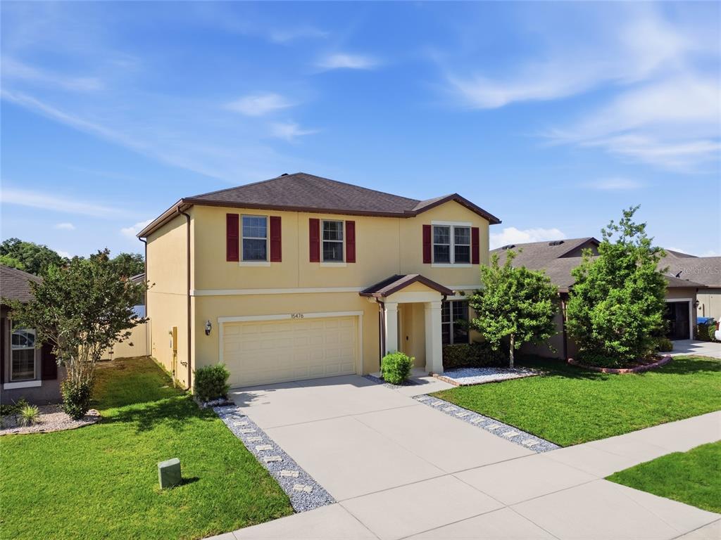 A two-story yellow house with a two-car garage and red shutters under a blue sky — 15476 STONE HOUSE DRIVE, BROOKSVILLE, FL