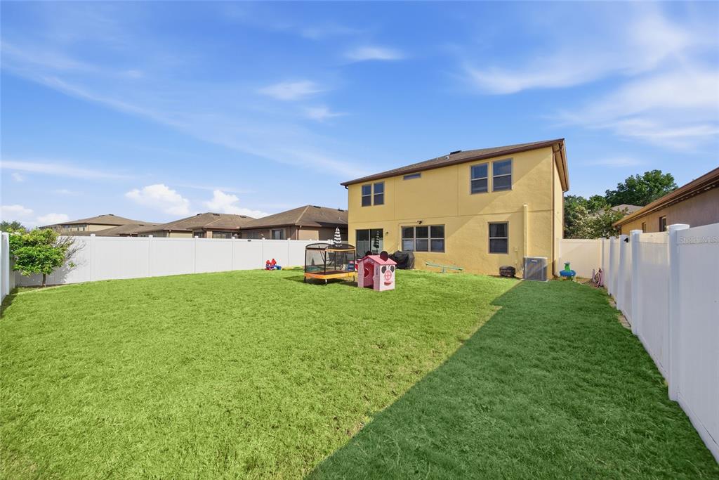 The backyard of a two-story yellow house features a lawn, white vinyl fencing, and toys — 15476 STONE HOUSE DRIVE, BROOKSVILLE, FL