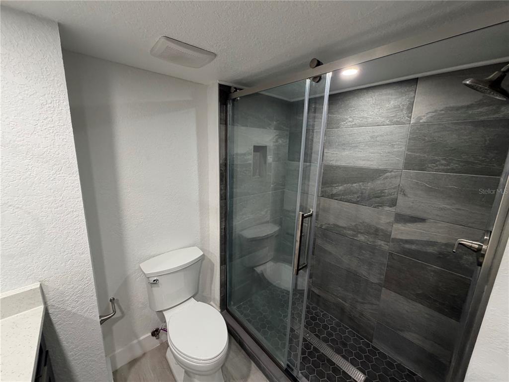 A bathroom features a white toilet next to a glass-enclosed shower with gray tiled walls — 13210 ARBOR ISLE DR UNIT 102, TEMPLE TERRACE, FL
