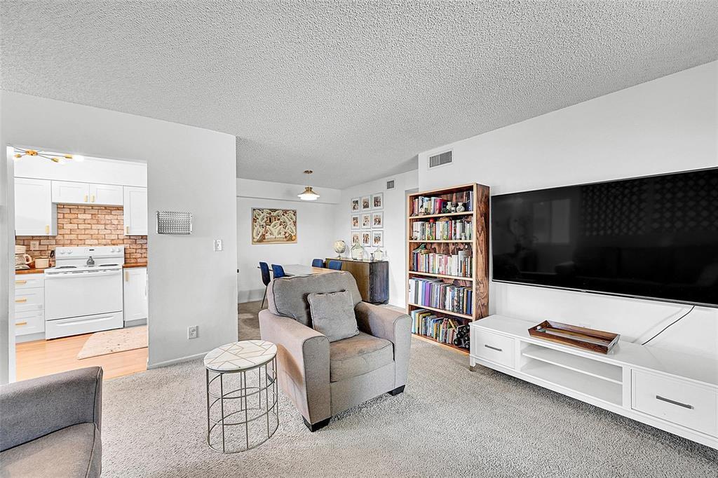 A living room with gray carpet, a television, a bookshelf, and an adjacent kitchen — 117 Gardens Drive # 204, Pompano Beach FL 33069, POMPANO BEACH, FL