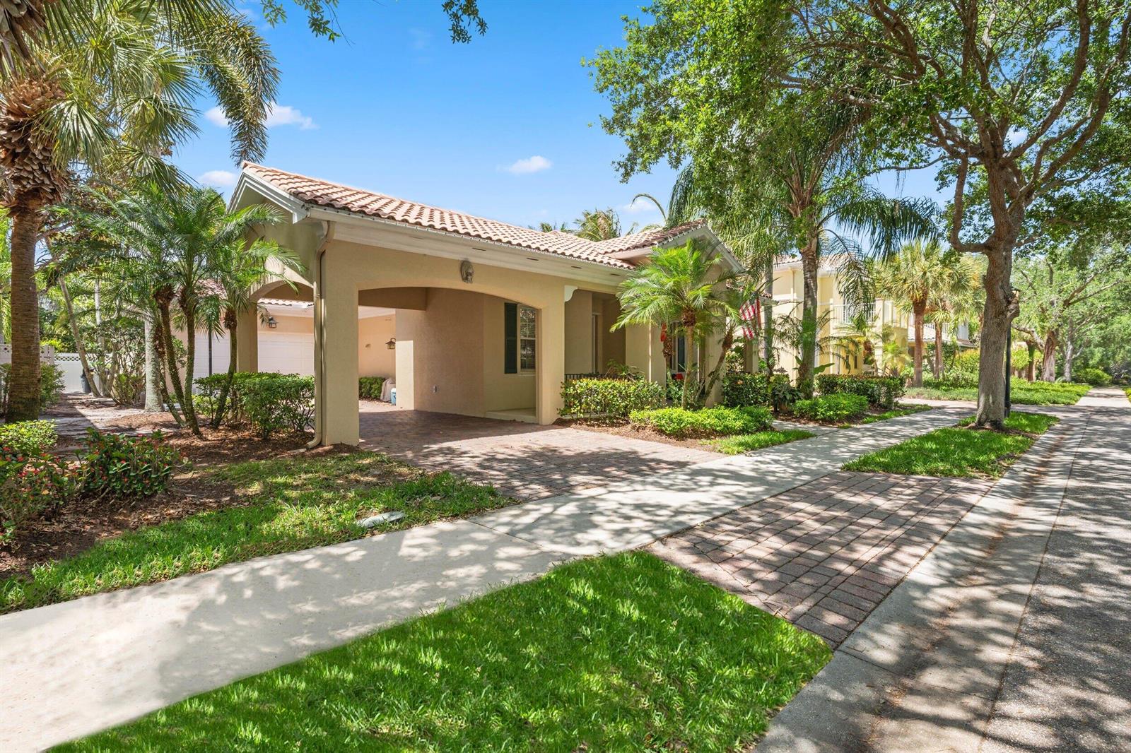 A tan single-story house with a covered entryway and a brick driveway surrounded by trees — 310 San Remo Drive, Jupiter FL 33458, JUPITER, FL