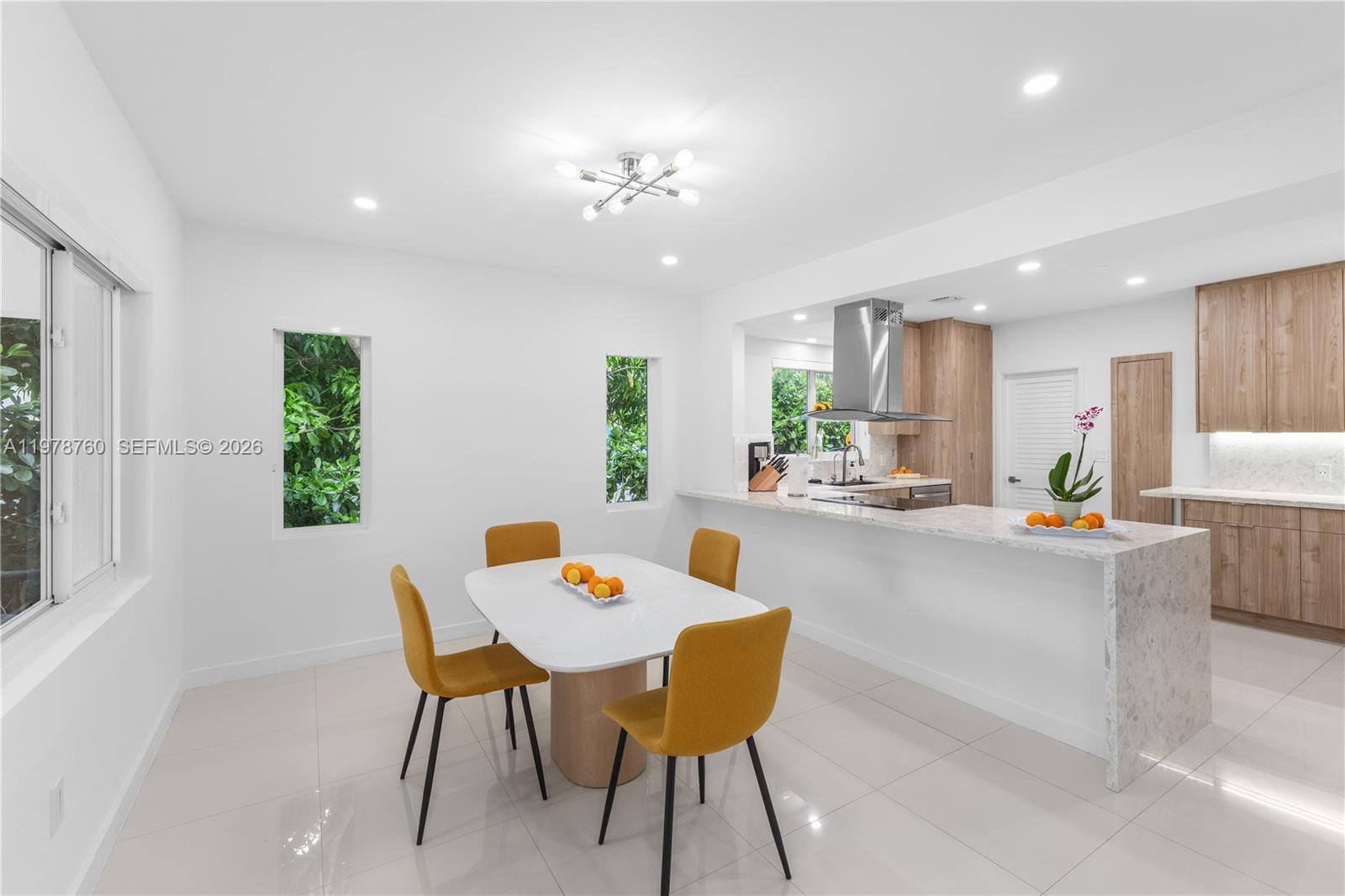 A dining area with yellow chairs sits adjacent to a modern kitchen with wooden cabinets — 741 NE 87th St, Miami FL 33138, MIAMI, FL