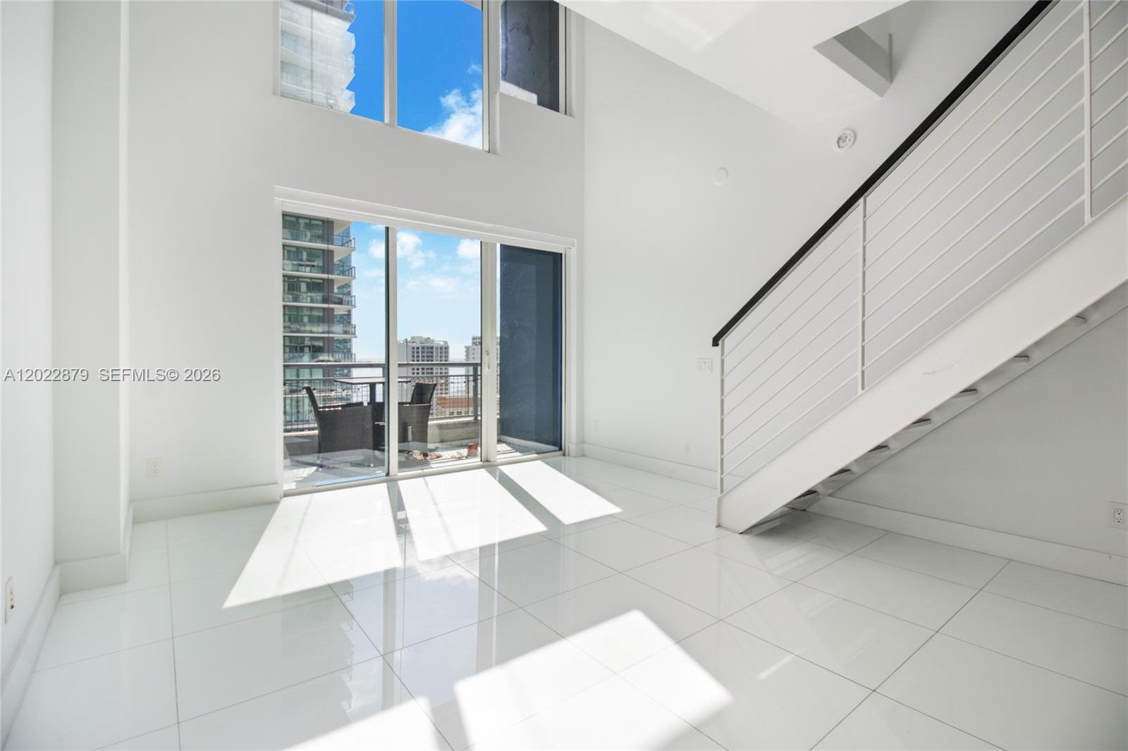 A bright, double-height living room features white tile flooring, a staircase, and a balcony view — 60 SW 13th St # 2208, Miami FL 33130, MIAMI, FL