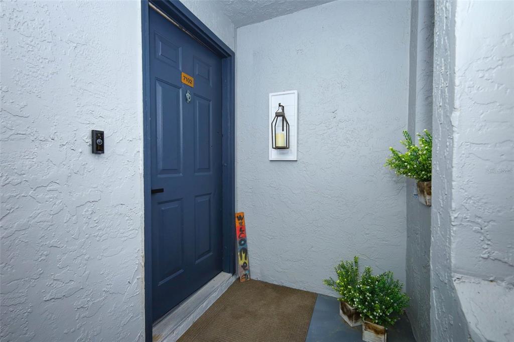 A blue front door with a welcome sign and wall decor sits in a stucco entryway — 7102 OLYMPIA COURT UNIT 7102, LONGWOOD, FL