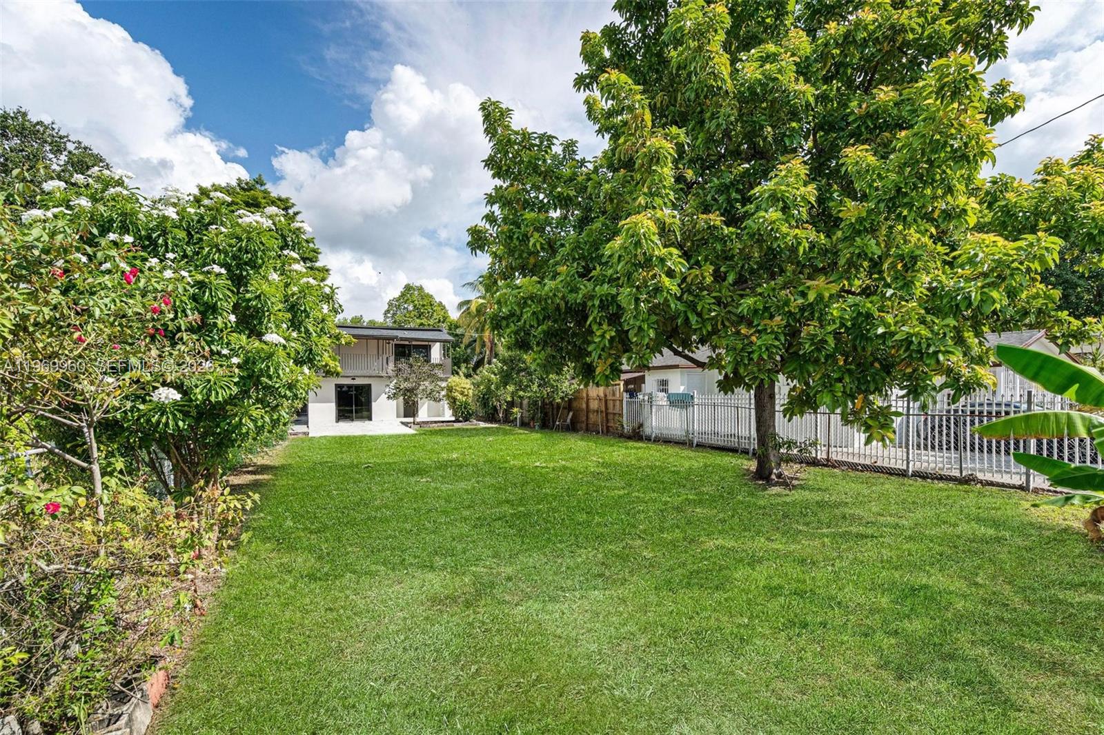 A grassy backyard features a large tree, a white fence, and a two-story house — 1523 NW 38th St, Miami FL 33142, MIAMI, FL