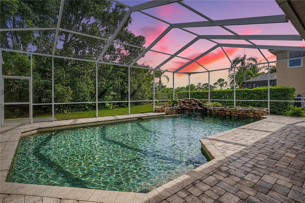 A screened-in swimming pool with a stone waterfall feature and paver patio at sunset — 1319 FORESTEDGE BOULEVARD, OLDSMAR, FL