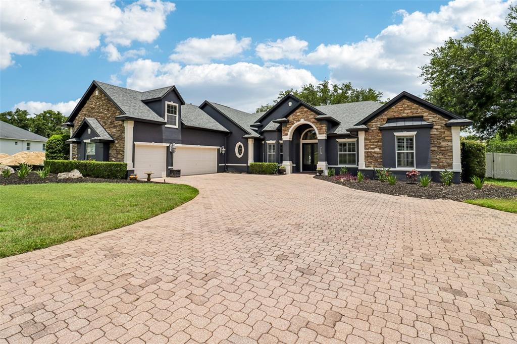 A dark-painted house with stone accents and a large paver driveway under a blue sky — 34028 MATTHEWS COVE, LEESBURG, FL