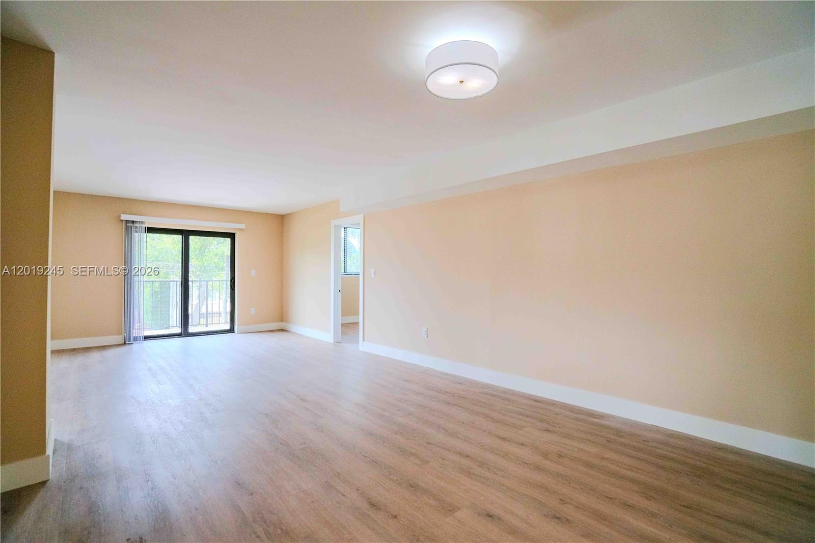 An empty living room features light-colored wood-look flooring, beige walls, and a sliding glass door — 6960 Rue Vendome # 201, Miami Beach FL 33141, MIAMI BEACH, FL
