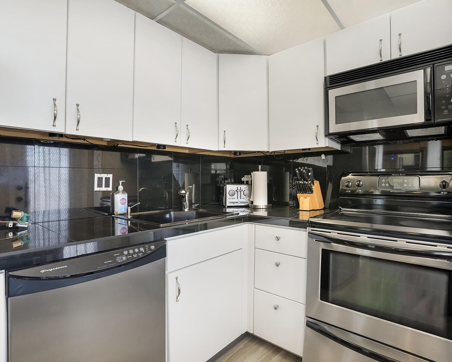 A kitchen features white cabinets, black countertops, stainless steel appliances, and a black tile backsplash — 2671 S Course Drive # 907, Pompano Beach FL 33069, POMPANO BEACH, FL