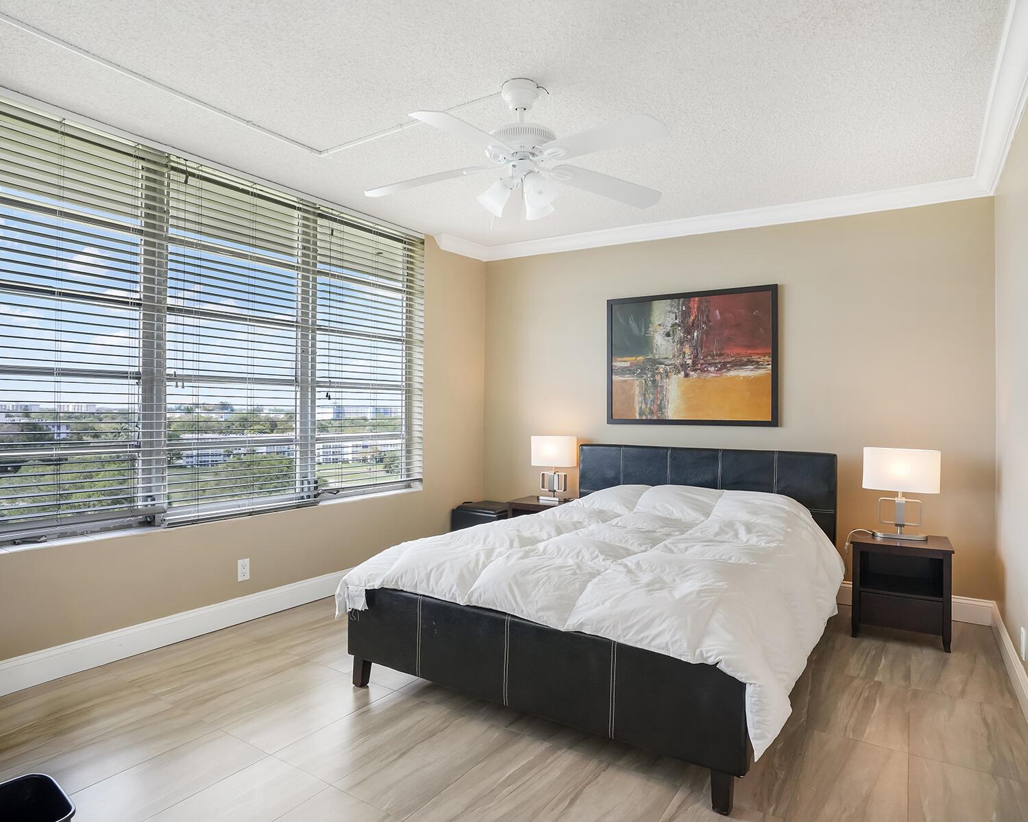 A bedroom features a black leather bed, two nightstands with lamps, and large windows with blinds — 2671 S Course Drive # 907, Pompano Beach FL 33069, POMPANO BEACH, FL