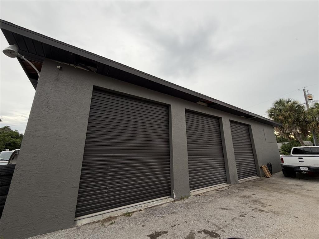 An exterior view of a gray building featuring three dark metal roll-up garage doors — 7805 CLARK MOODY BOULEVARD, PORT RICHEY, FL