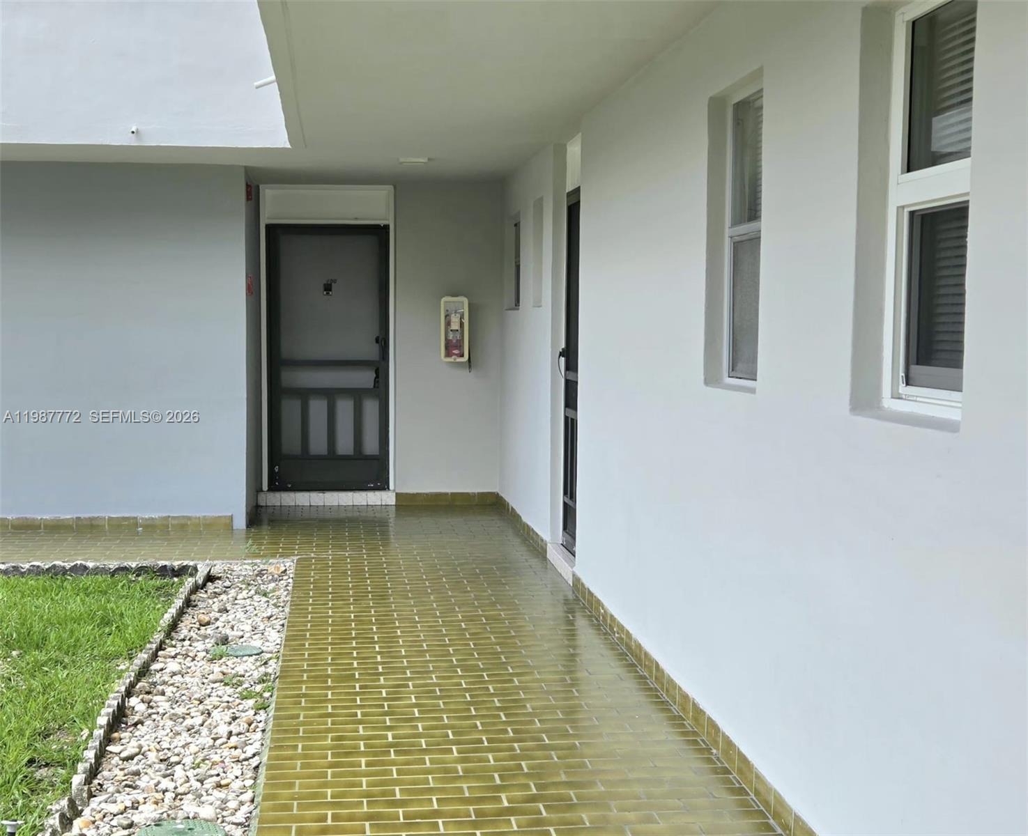 A covered walkway with green tiled flooring leads to two doors along a white wall — 1700 NE 191st St # 100, Miami FL 33179, MIAMI, FL