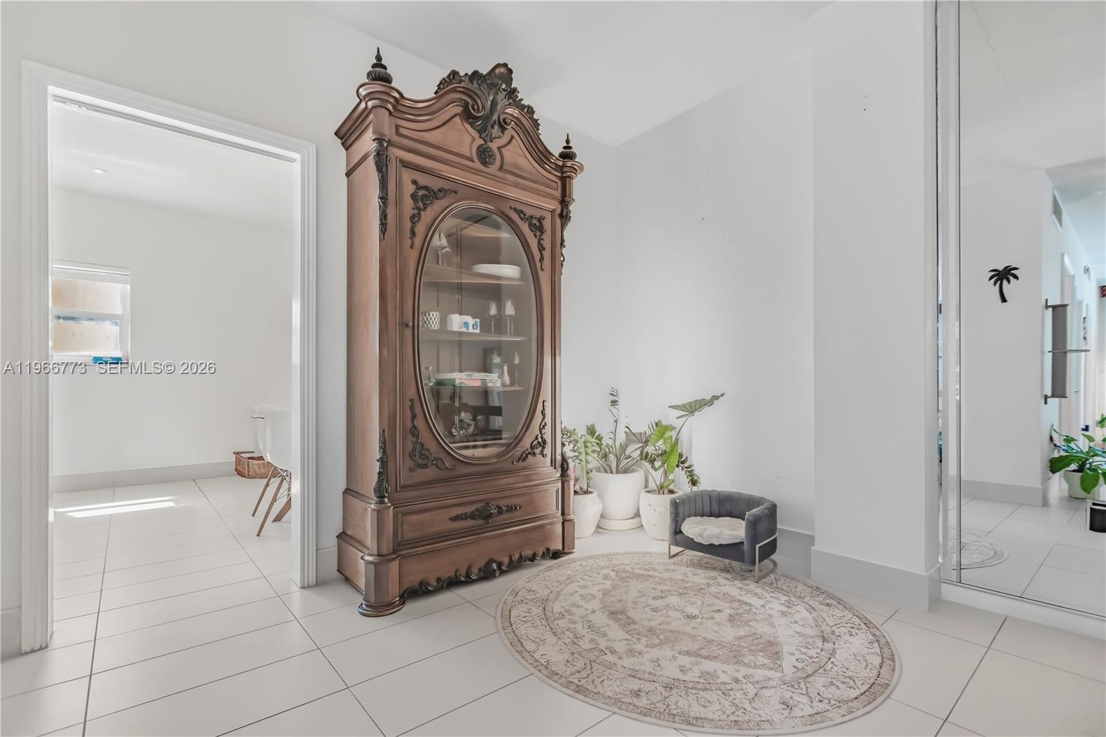 A large ornate wooden display cabinet stands in a white-tiled room with potted plants — 5001 Collins Ave # 1K, Miami Beach FL 33140, MIAMI BEACH, FL