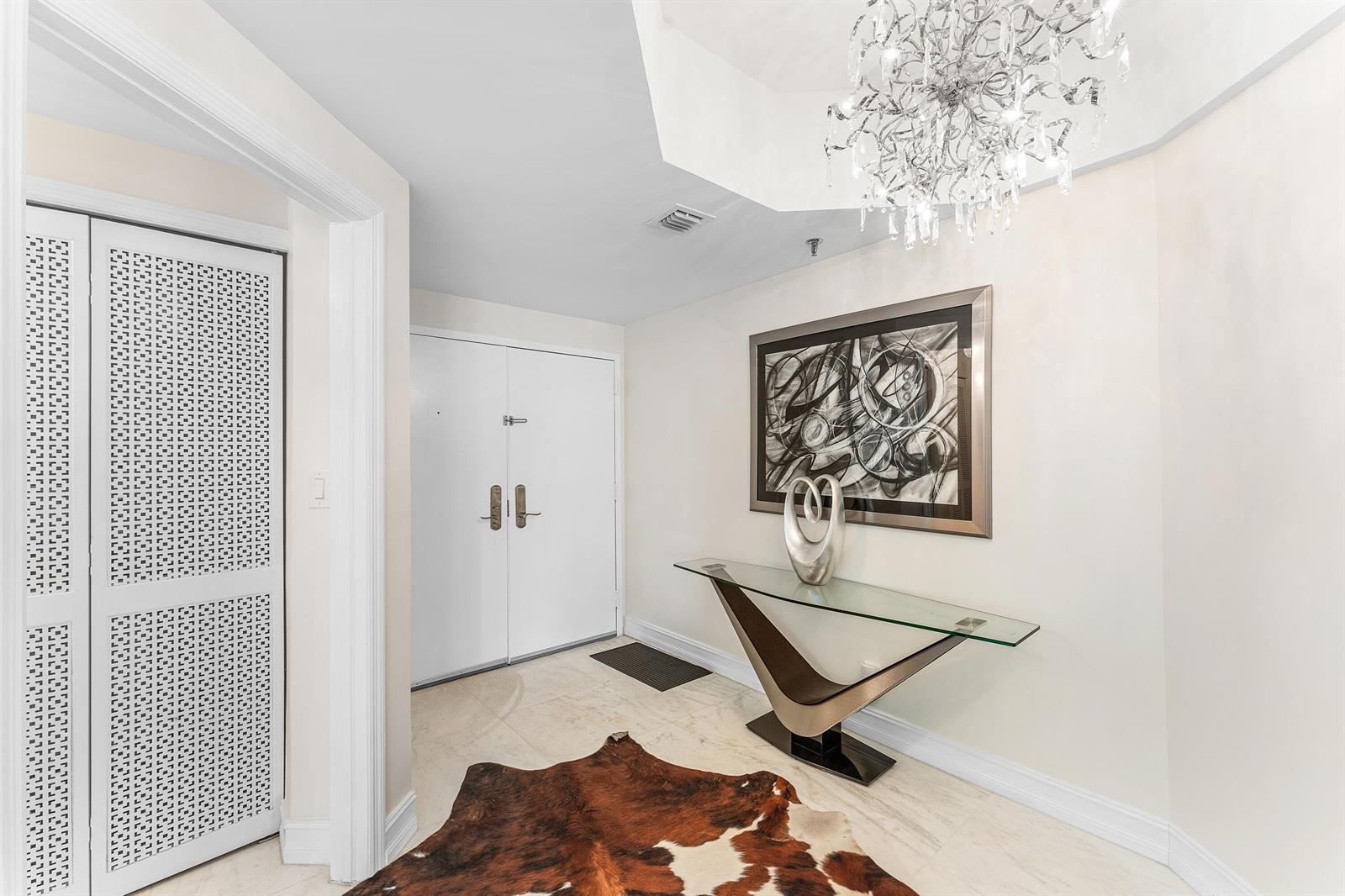 A foyer features a glass console table, a cowhide rug, and a crystal chandelier — 3120 S Ocean Boulevard # 202, Palm Beach FL 33480, PALM BEACH, FL