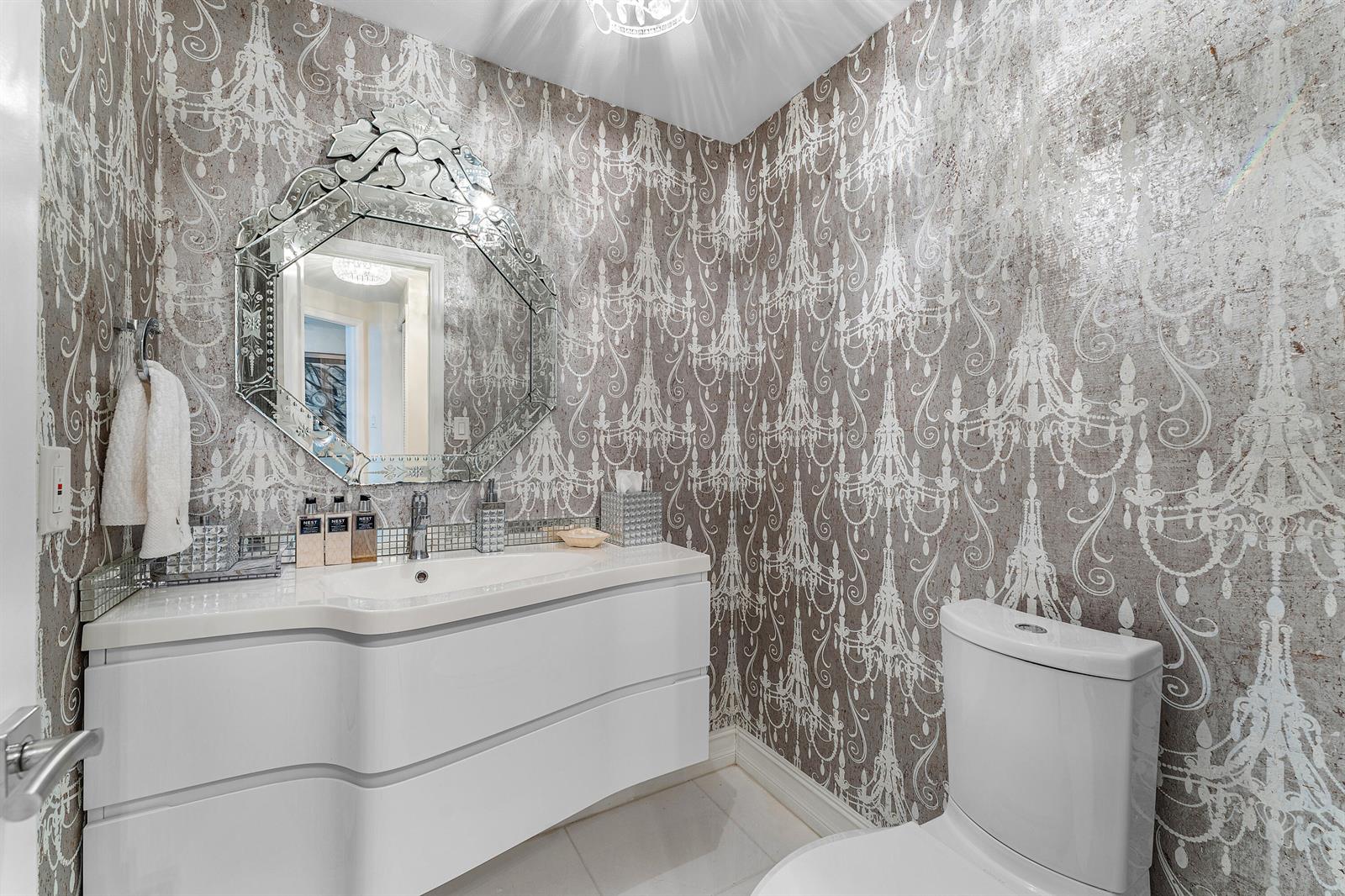 A powder room features chandelier-patterned wallpaper, a modern white vanity, and an ornate Venetian mirror — 3120 S Ocean Boulevard # 202, Palm Beach FL 33480, PALM BEACH, FL