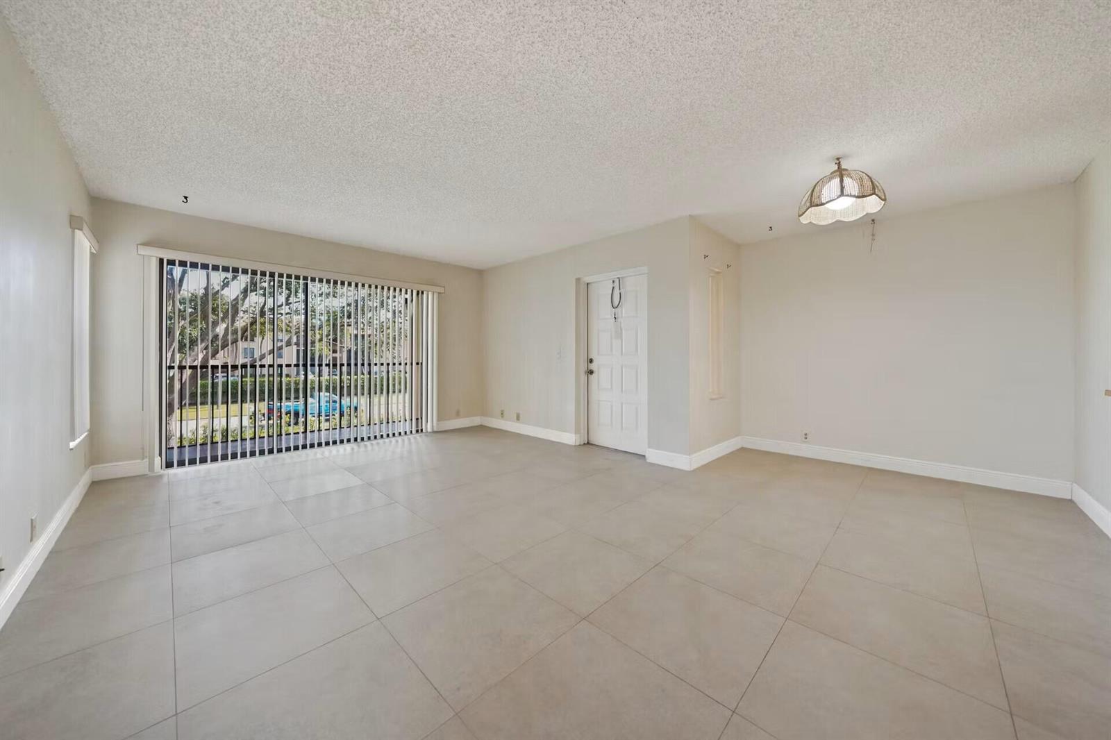 An empty living room with large beige floor tiles, a popcorn ceiling, and vertical blinds — 802 Sky Pine Way # E2, FL 33415, FL