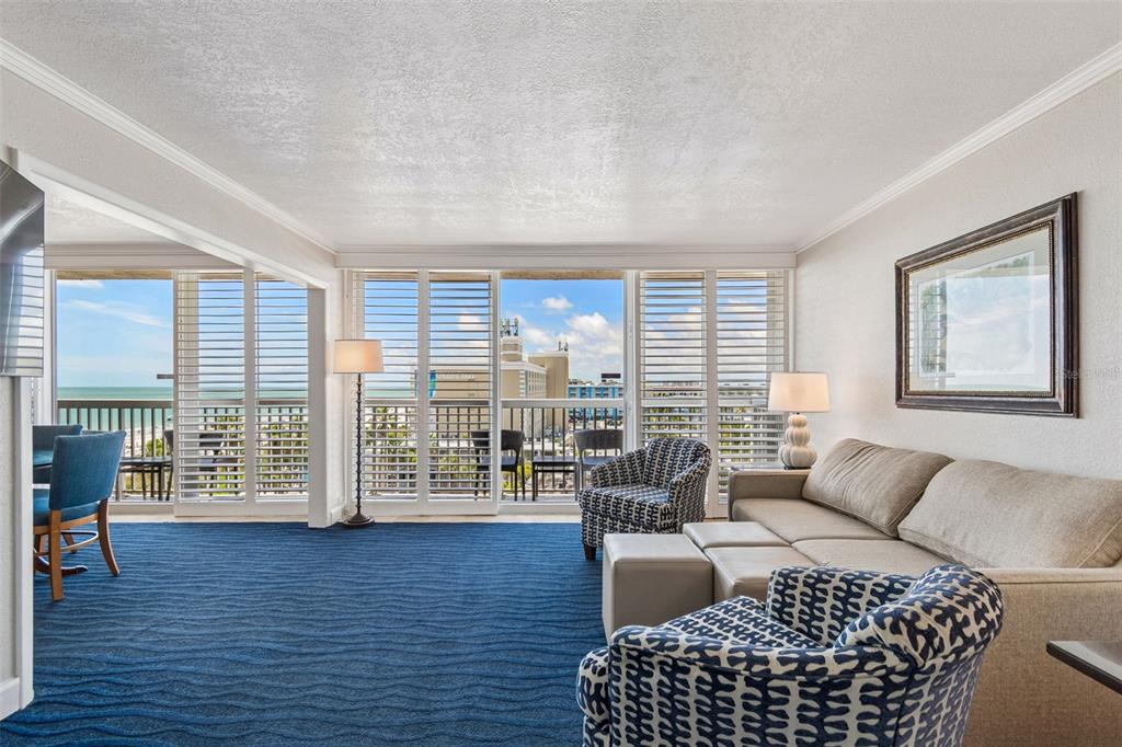 A living room with blue carpet, patterned armchairs, a beige sofa, and floor-to-ceiling balcony shutters — 5500 GULF BOULEVARD UNIT 7230, ST PETE BEACH, FL